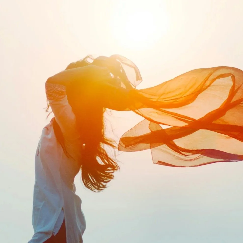 A woman with long hair standing against a bright sun, holding an orange translucent scarf that is flowing in the wind.