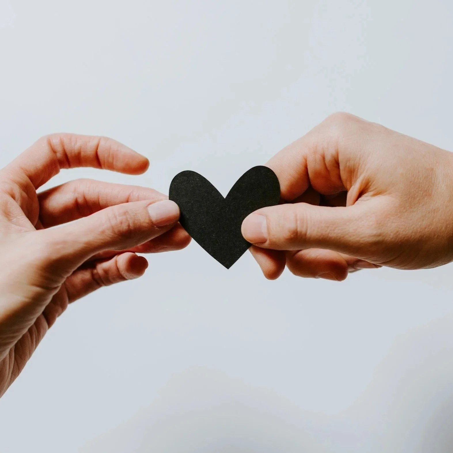 Two hands exchanging a black paper heart against a plain white background.