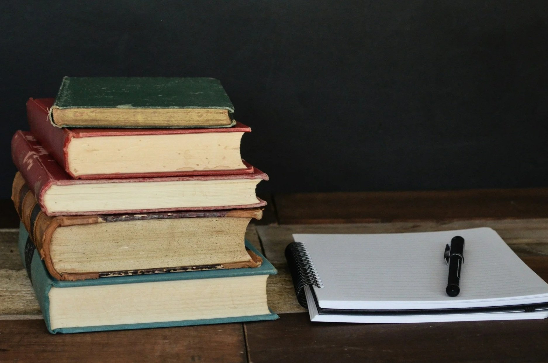 A stack of old hardcover books with a spiral notepad and pen on a wooden surface.