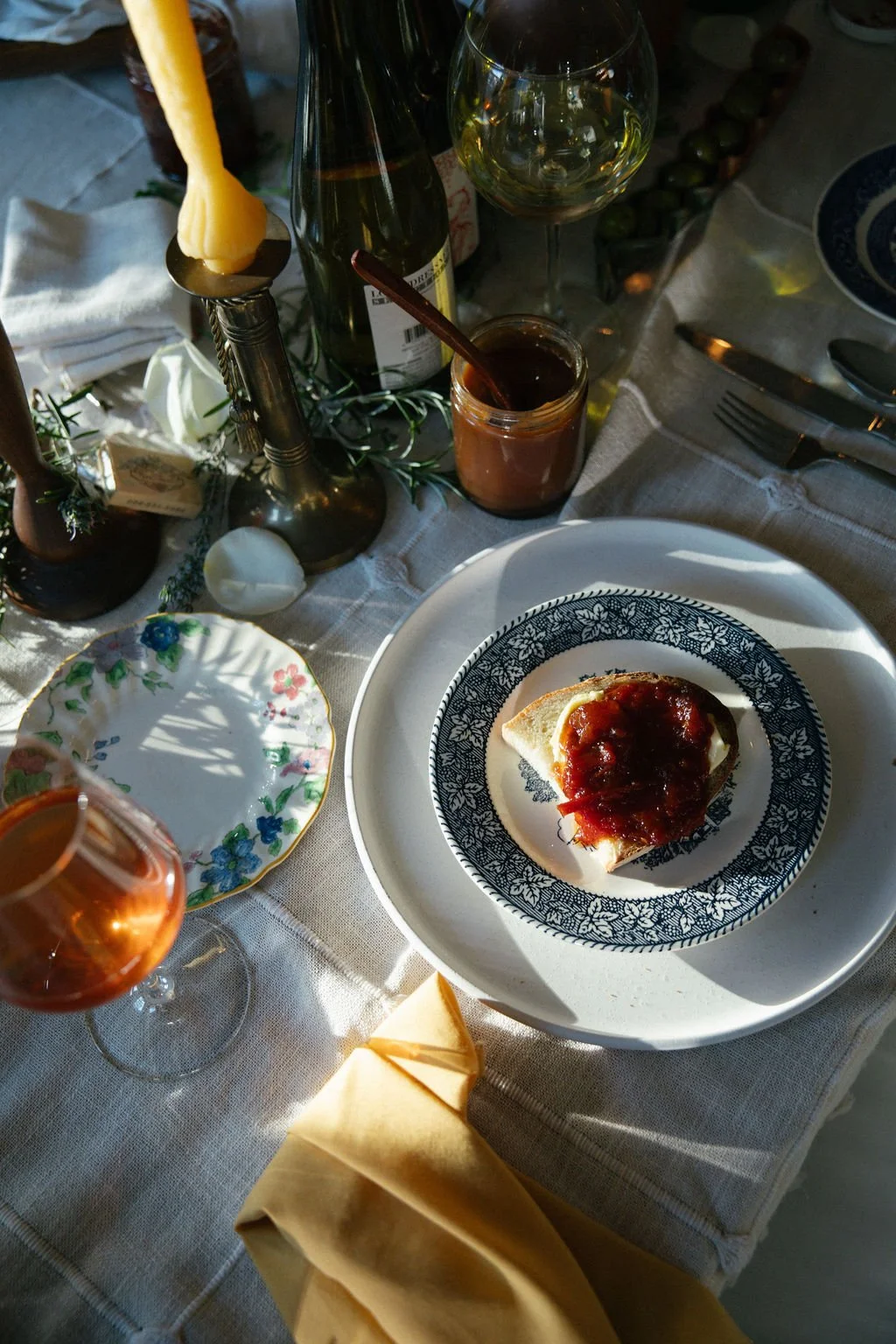 A table setting with a slice of bread topped with butter and tomato sauce, a glass of rosé wine, a jar of jam, a candle, and various dishes and utensils on a white tablecloth.