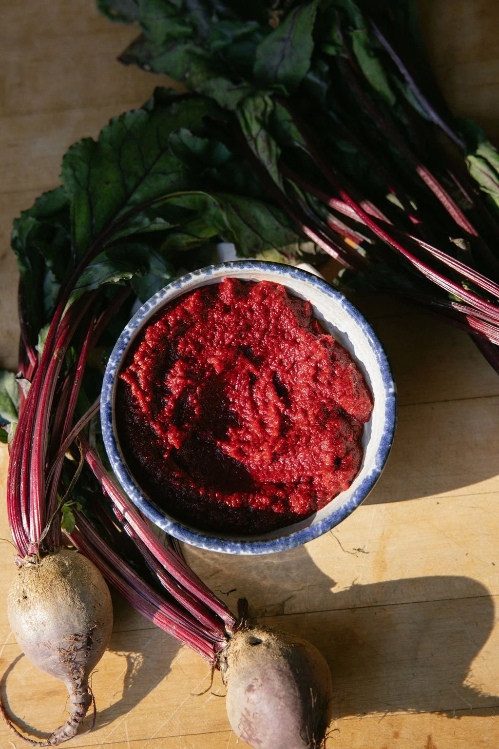 Fresh beets with leafy greens surrounding a bowl of beetroot hummus on a wooden surface.