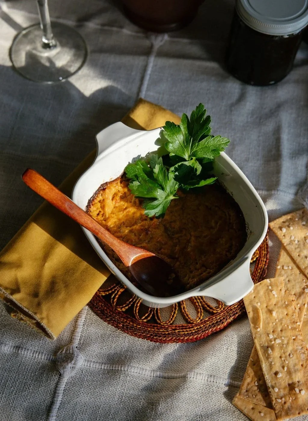Baked dish garnished with parsley at a dining table, with saltine crackers beside it, and a glass of red wine and a jar in the background.