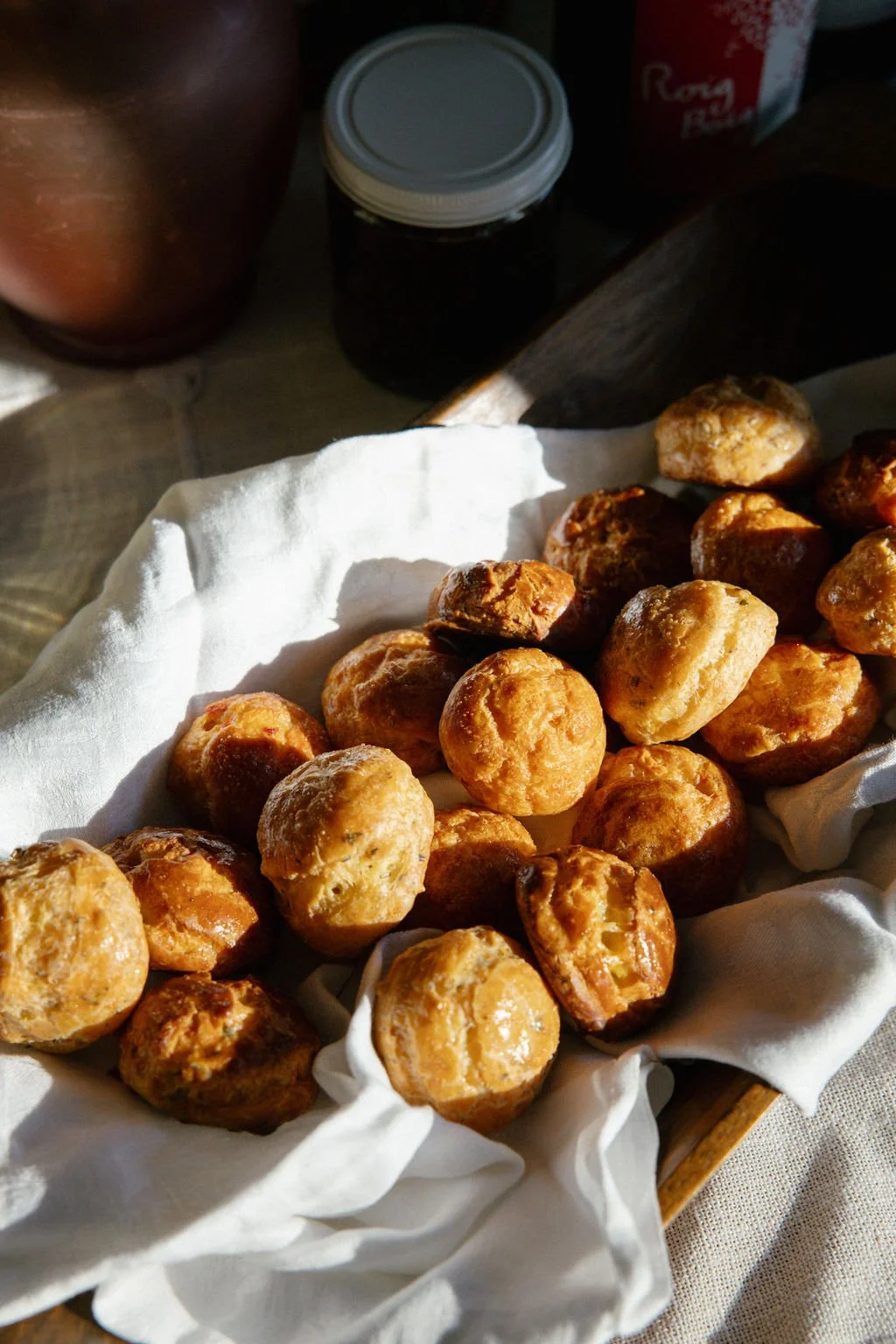 A basket of freshly baked, golden-brown bread rolls on a white cloth, with jars and a cup in the background.