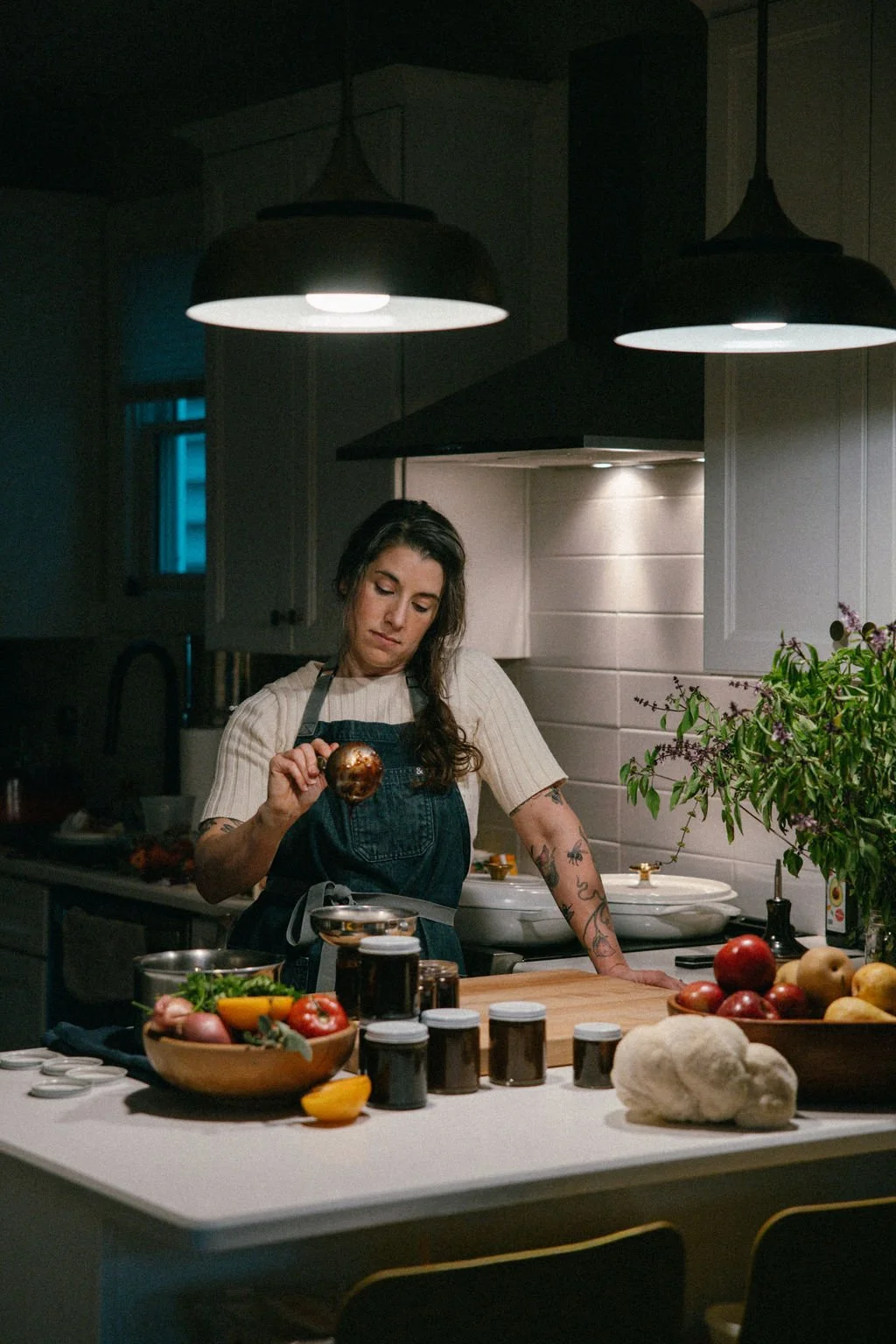 A woman with tattoos and dark hair in a kitchen, wearing a white shirt and denim apron, is holding a garlic bulb over a bowl, surrounded by fresh vegetables, jars of spices, and a potted plant, under warm overhead lighting.