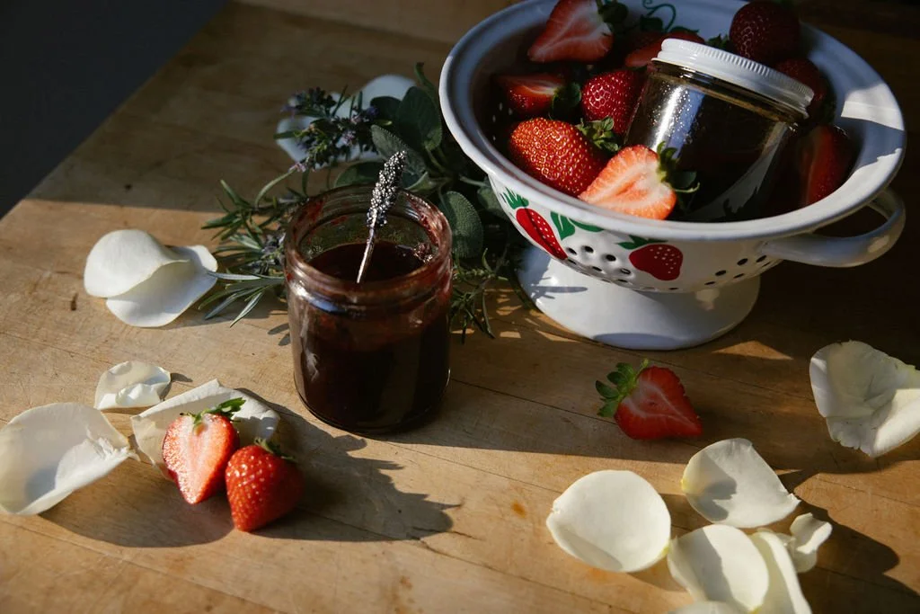 Strawberries in a white bowl and on a wooden table, surrounded by white rose petals, with a small jar of jam and a jar of strawberry preserves, under natural sunlight.