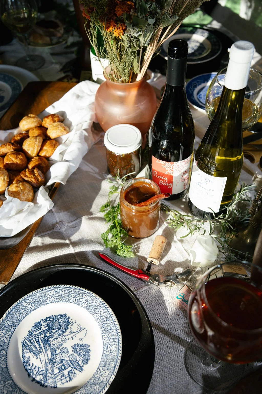 A table set for a meal with bread rolls, wine bottles, jars of jam or preserves, glasses of wine, a floral centerpiece, and a decorative plate.