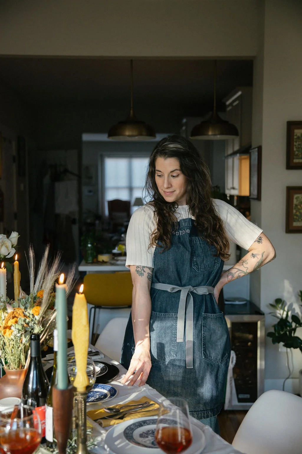 Woman with long dark hair and tattoos on her arms standing next to a decorated dining table in a home, wearing a denim apron over a white shirt, during daytime.
