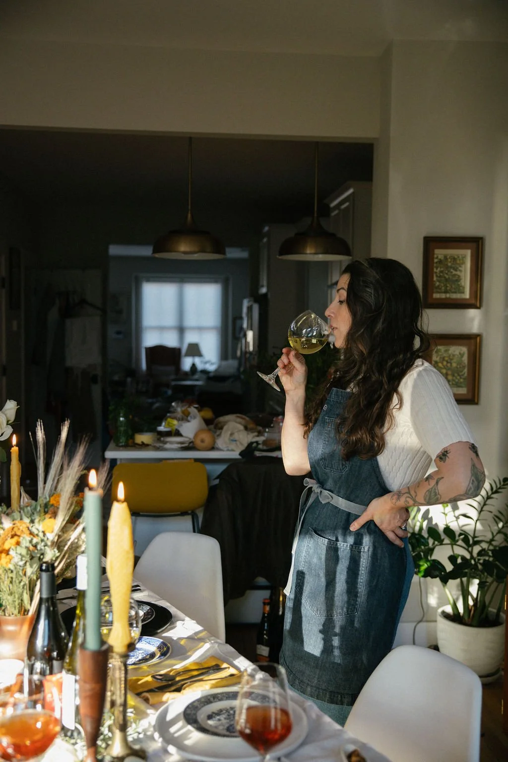 A woman with tattoos and dark curly hair, wearing a white shirt and denim apron, drinks white wine in a dining room with a decorated table.