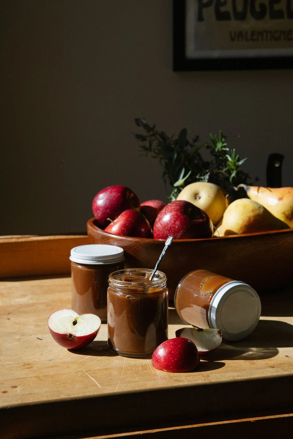A wooden table with jars of apple butter, some apples, and a bowl of mixed fruits in the background in a dimly lit room.