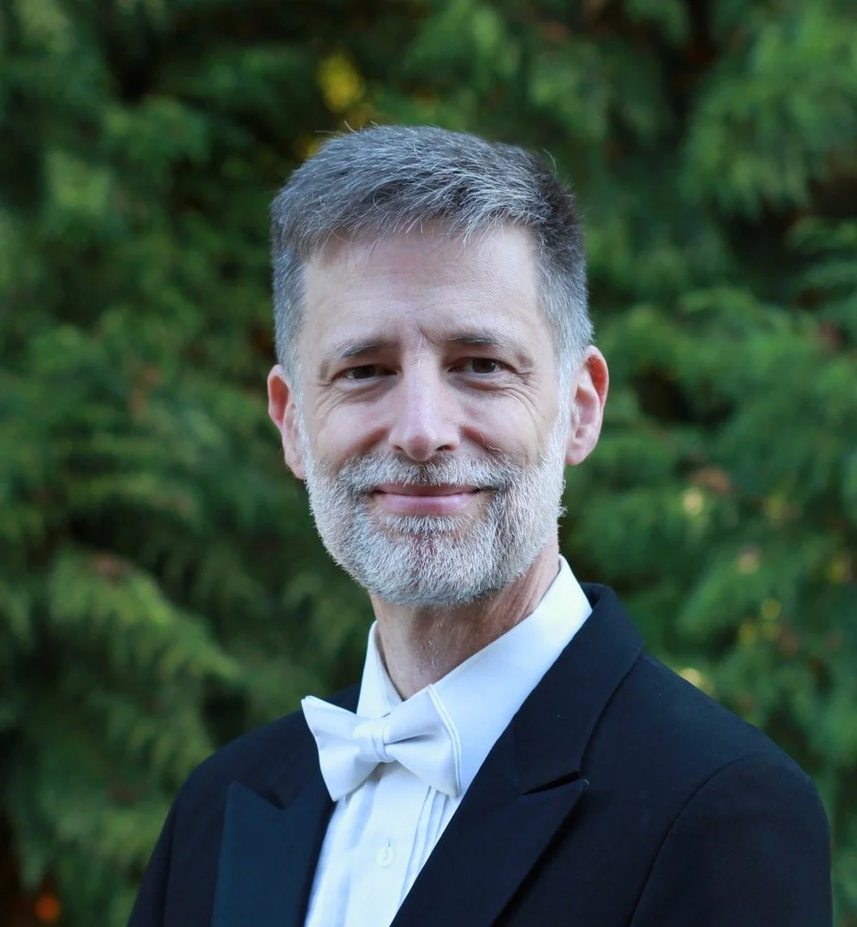 A mature man with gray hair and a beard, dressed in a tuxedo with a white bow tie, standing outdoors in front of greenery, smiling at the camera.