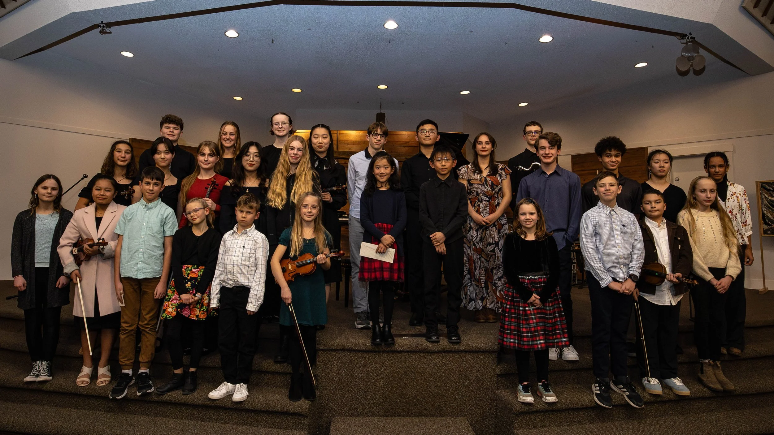 Group of children and teens standing on stage in a concert hall, some holding violins, all facing forward and smiling.