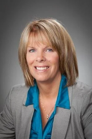 Professional woman with blonde hair, wearing a gray blazer and blue blouse, smiling against a neutral background.