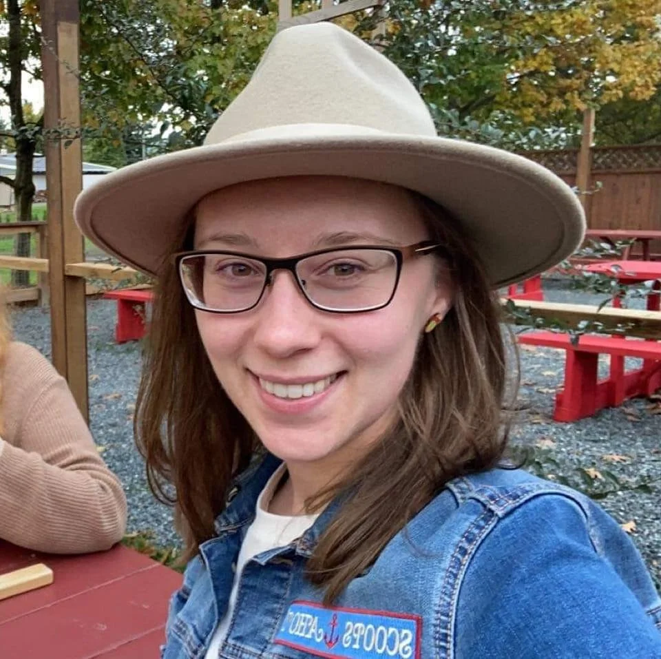 A young woman smiling outdoors, wearing a tan wide-brim hat, glasses, a denim jacket with a 'SCOOPS A' patch, and earrings. There are outdoor tables and trees in the background.
