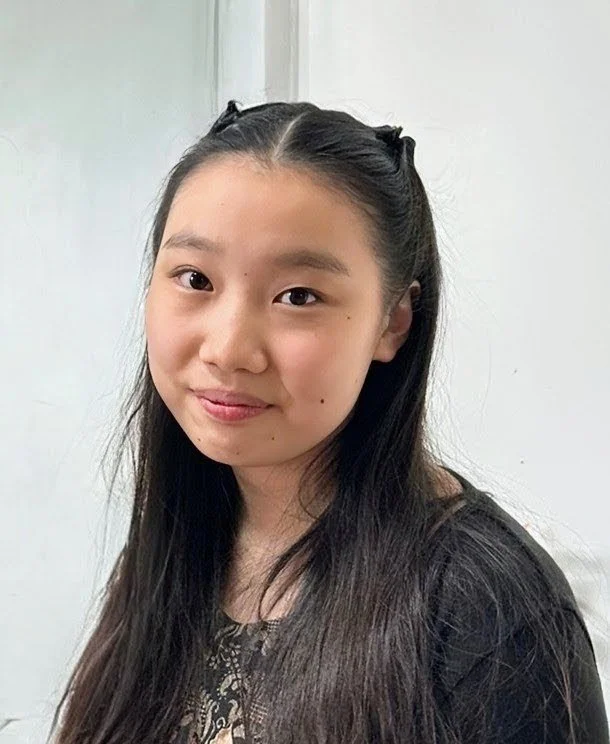 A young Asian girl with long black hair, smiling softly, wearing a black top with a patterned neckline, standing in front of a plain white wall.