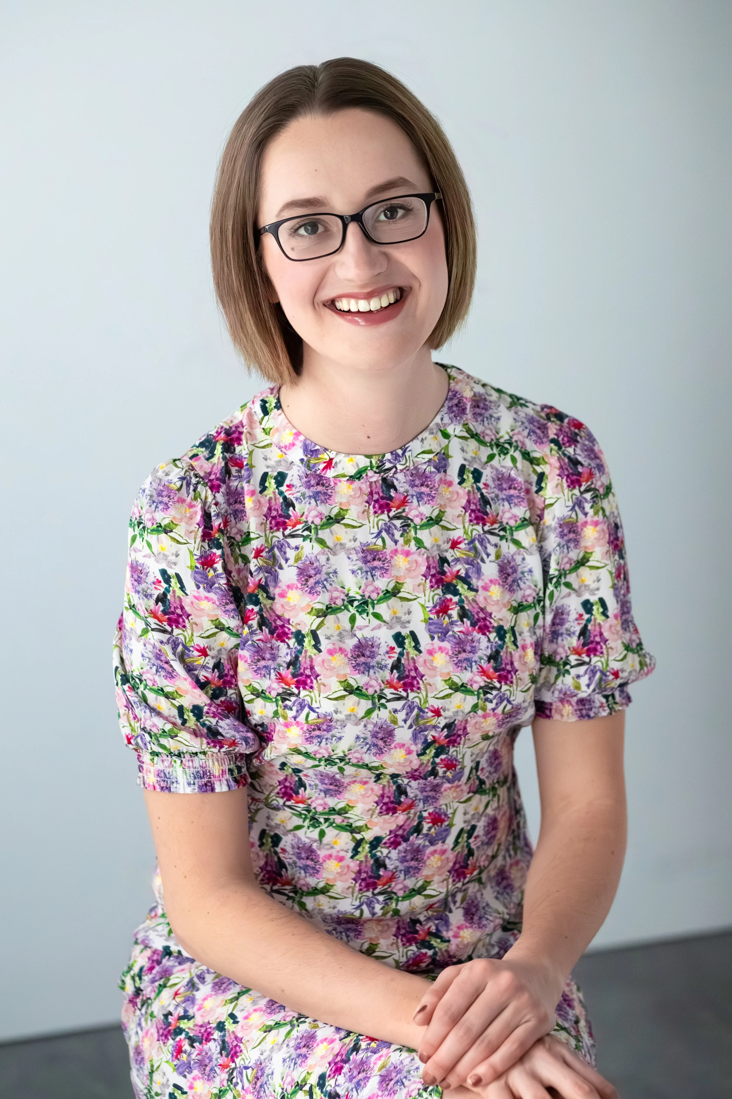 A young woman with shoulder-length brown hair, wearing glasses and a floral dress, smiling while seated against a plain light-colored background.