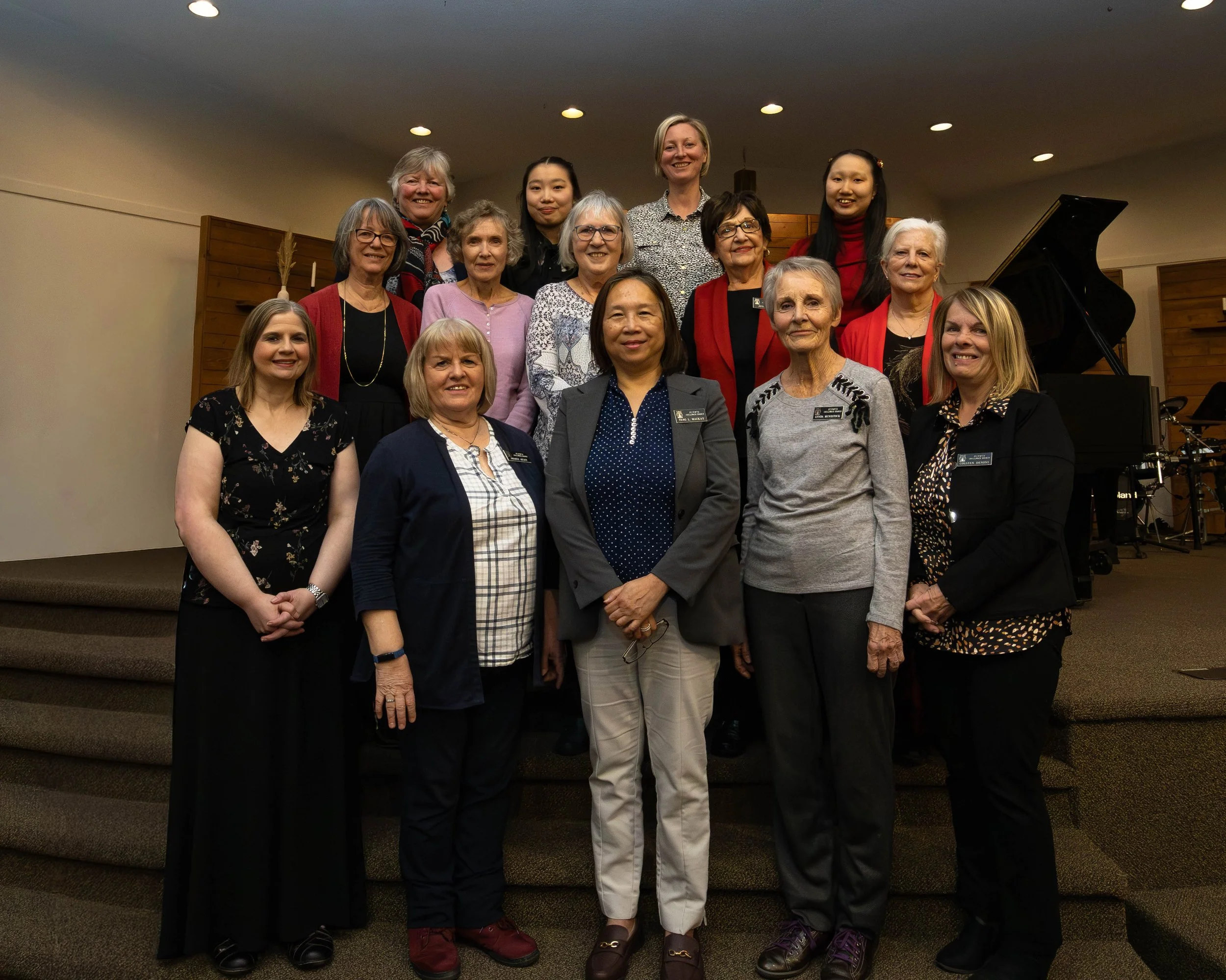 A group of 15 women standing together on stage in a church or auditorium, with a piano in the background, smiling at the camera.