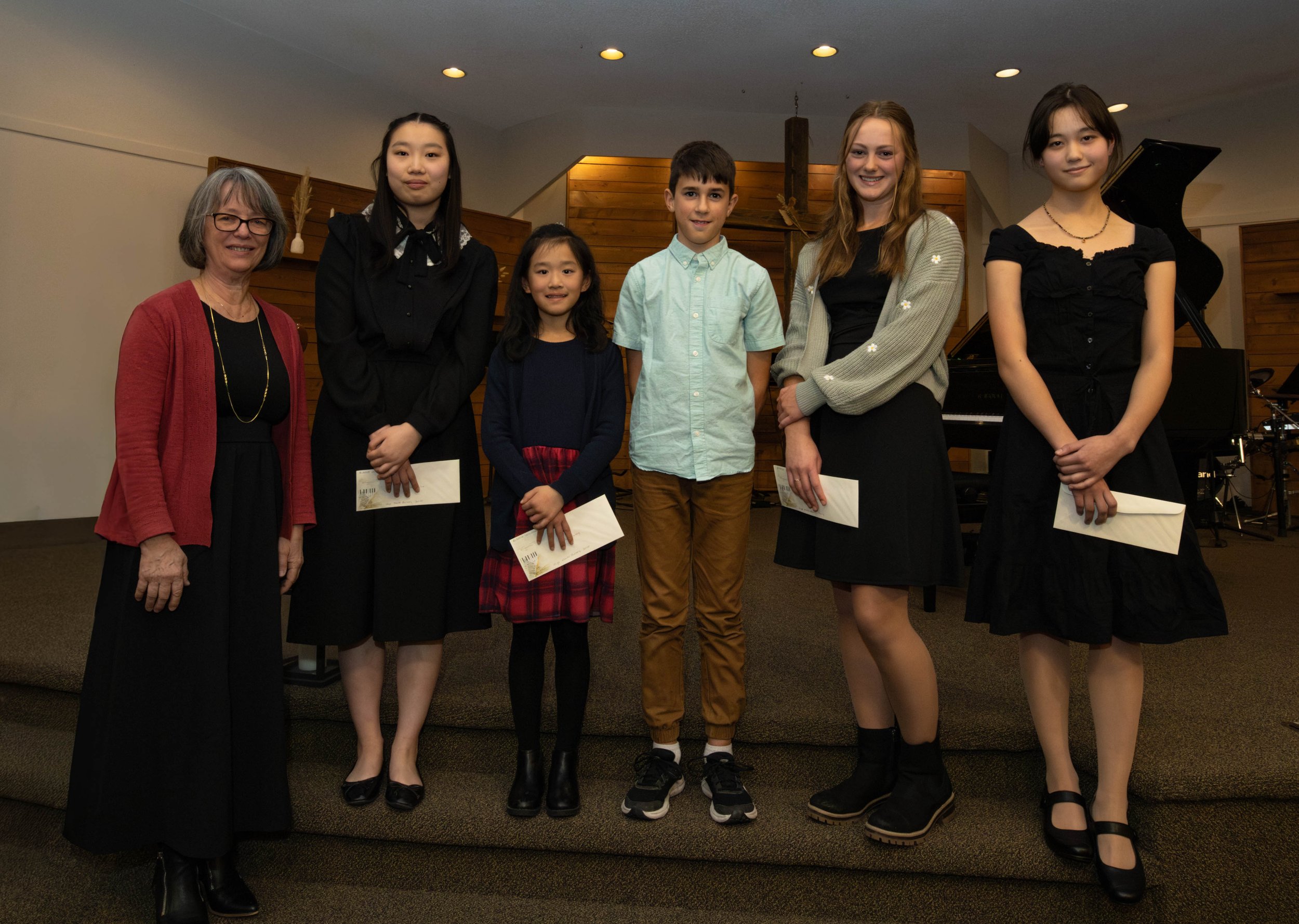 A group of six people, five young students and one older woman, standing on a stage indoor. The students are holding envelopes and appear to have been awarded certificates. The setting suggests a musical or awards ceremony, with a grand piano visible