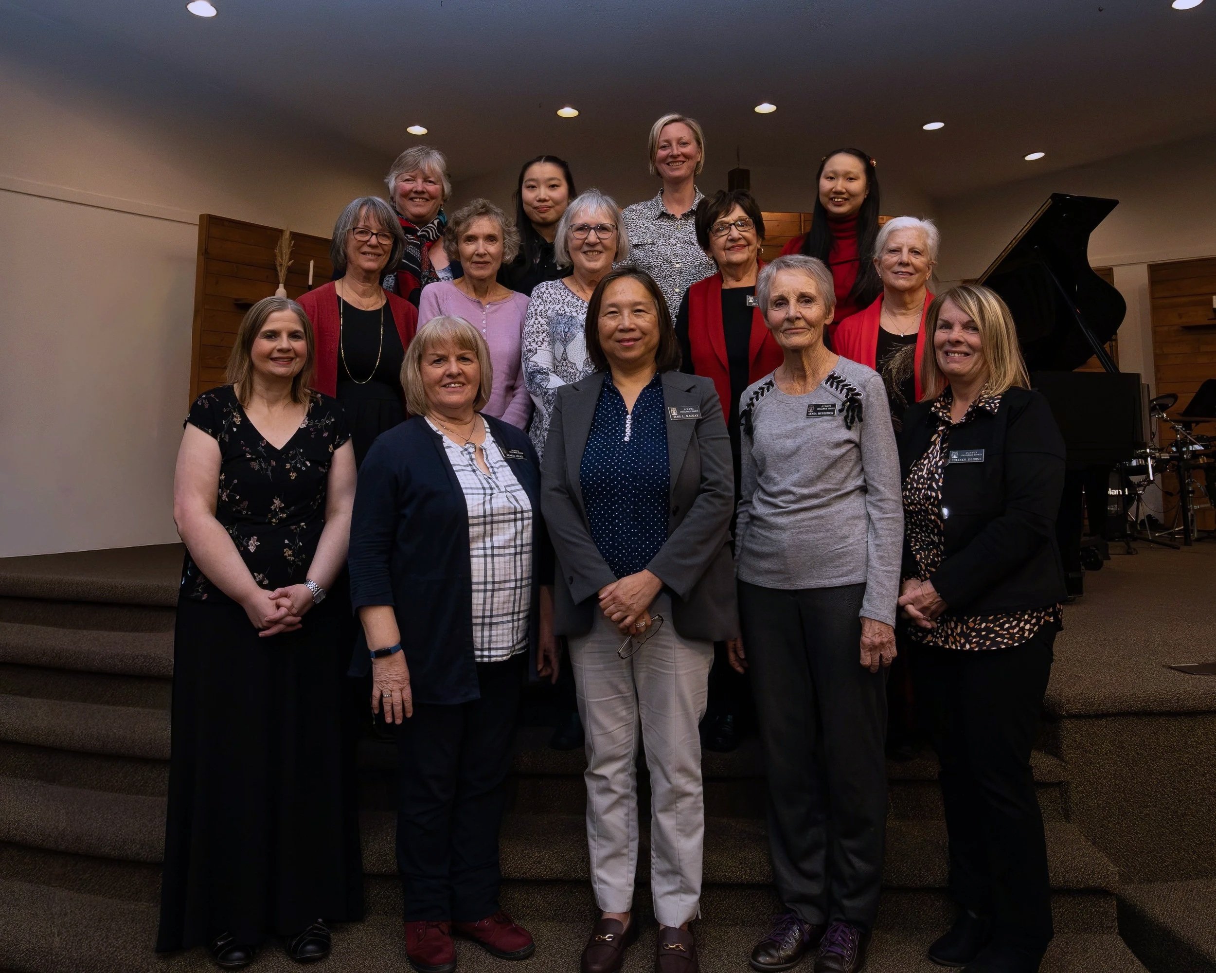 Group of 15 women standing on stage in front of a grand piano, smiling for a photo.