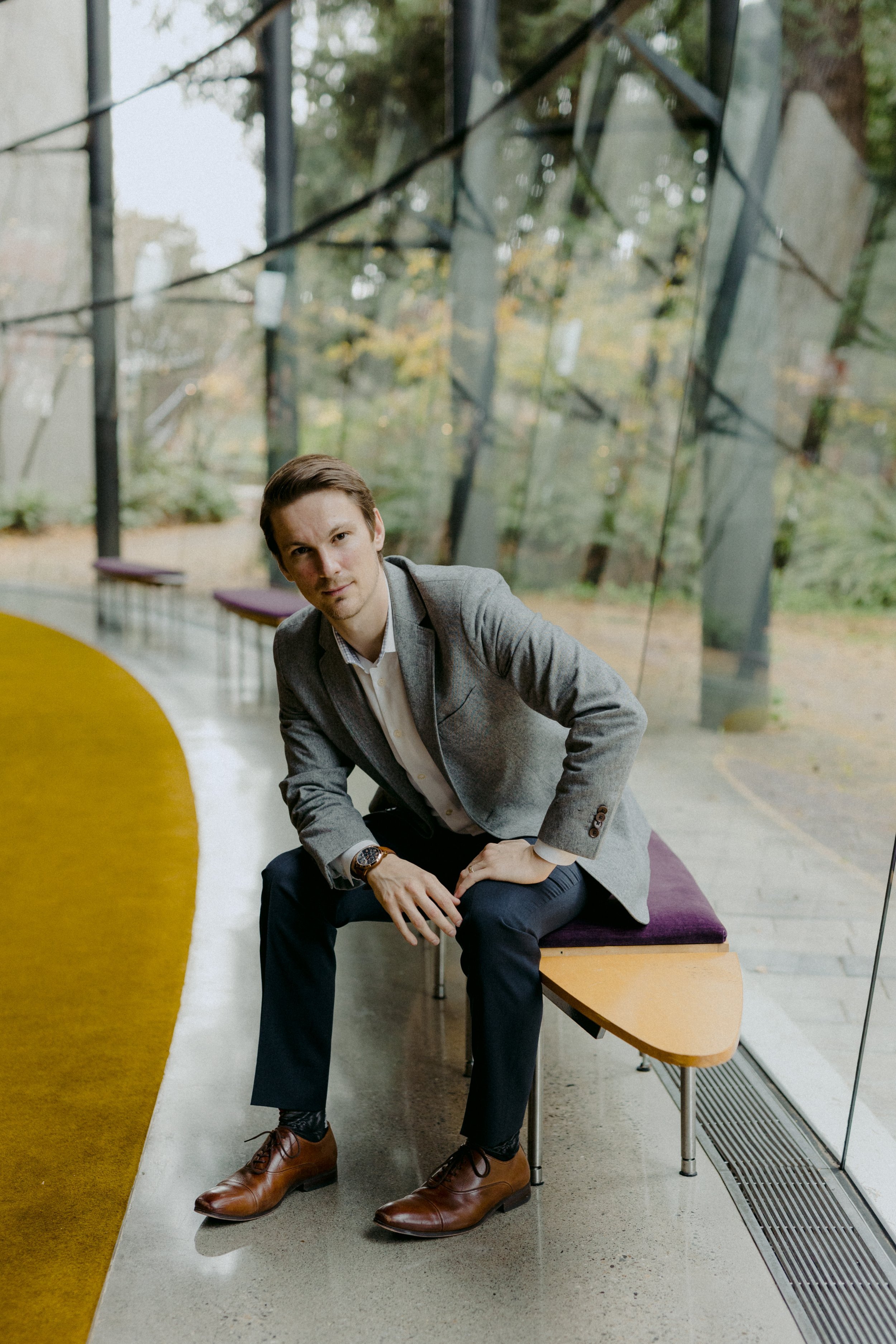 A man in a gray blazer and dark pants, sitting on a bench with a purple cushion, inside a modern glass building with a view of trees outside.