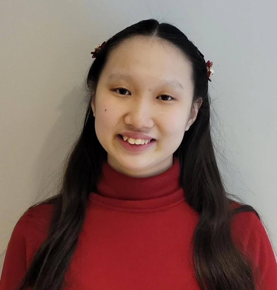 A young woman smiling, wearing a red turtleneck sweater, with long dark hair and hair accessories, standing against a plain background.