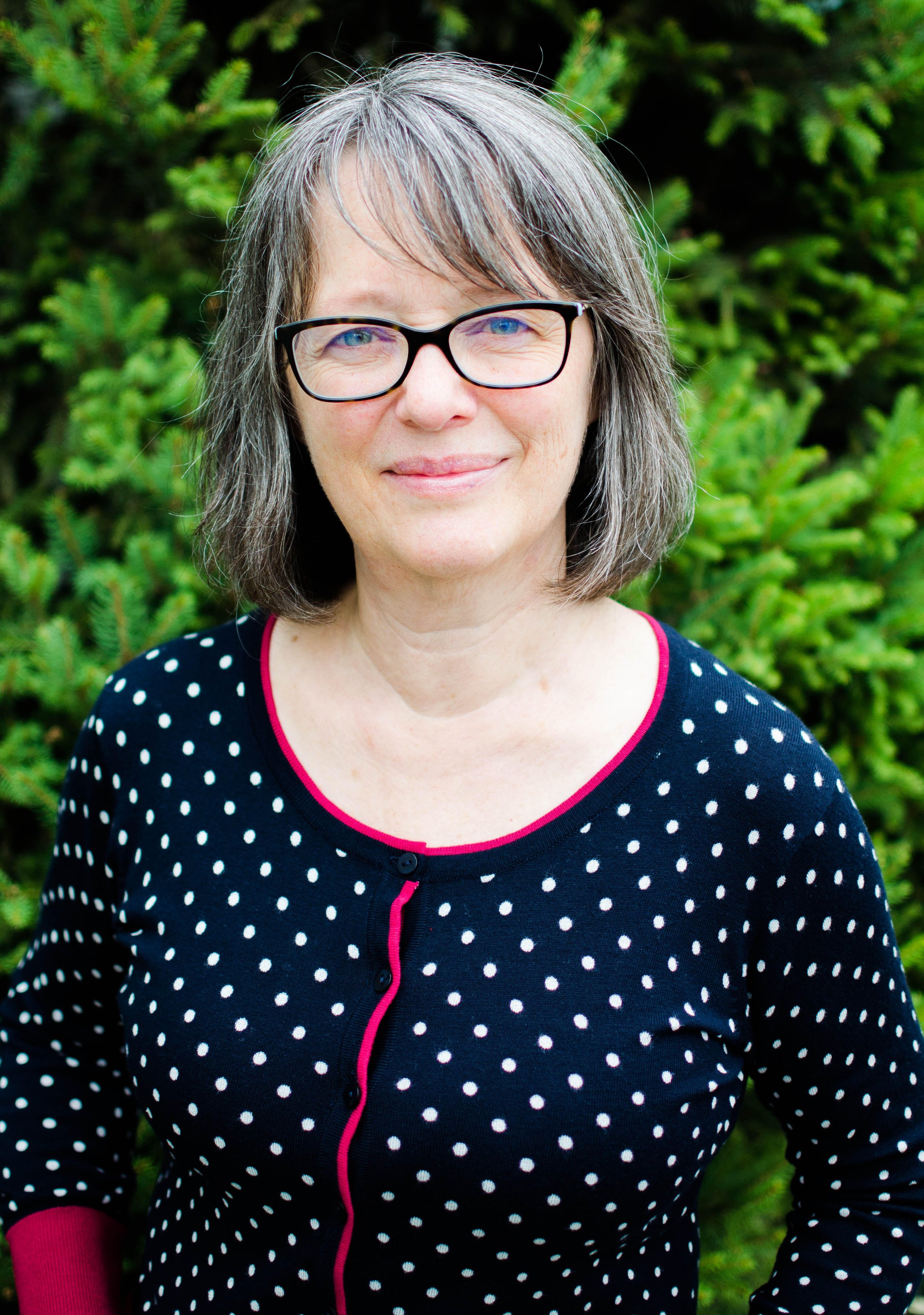 A middle-aged woman with shoulder-length gray hair and black glasses smiling outdoors with green foliage in the background, wearing a black polka dot sweater with pink trim.