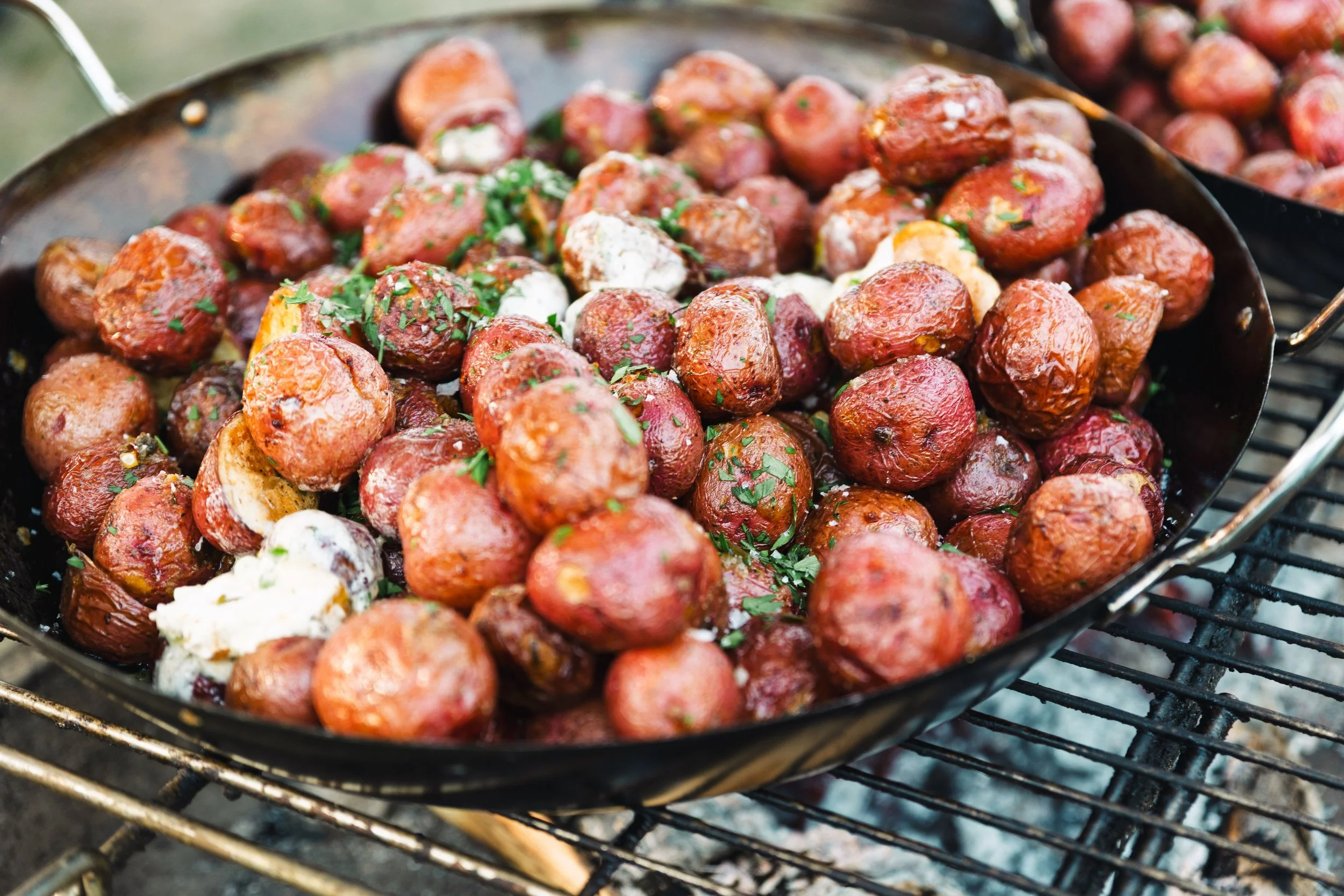Cooking small red potatoes in a black skillet over an outdoor grill, garnished with chopped herbs and dollops of sour cream.