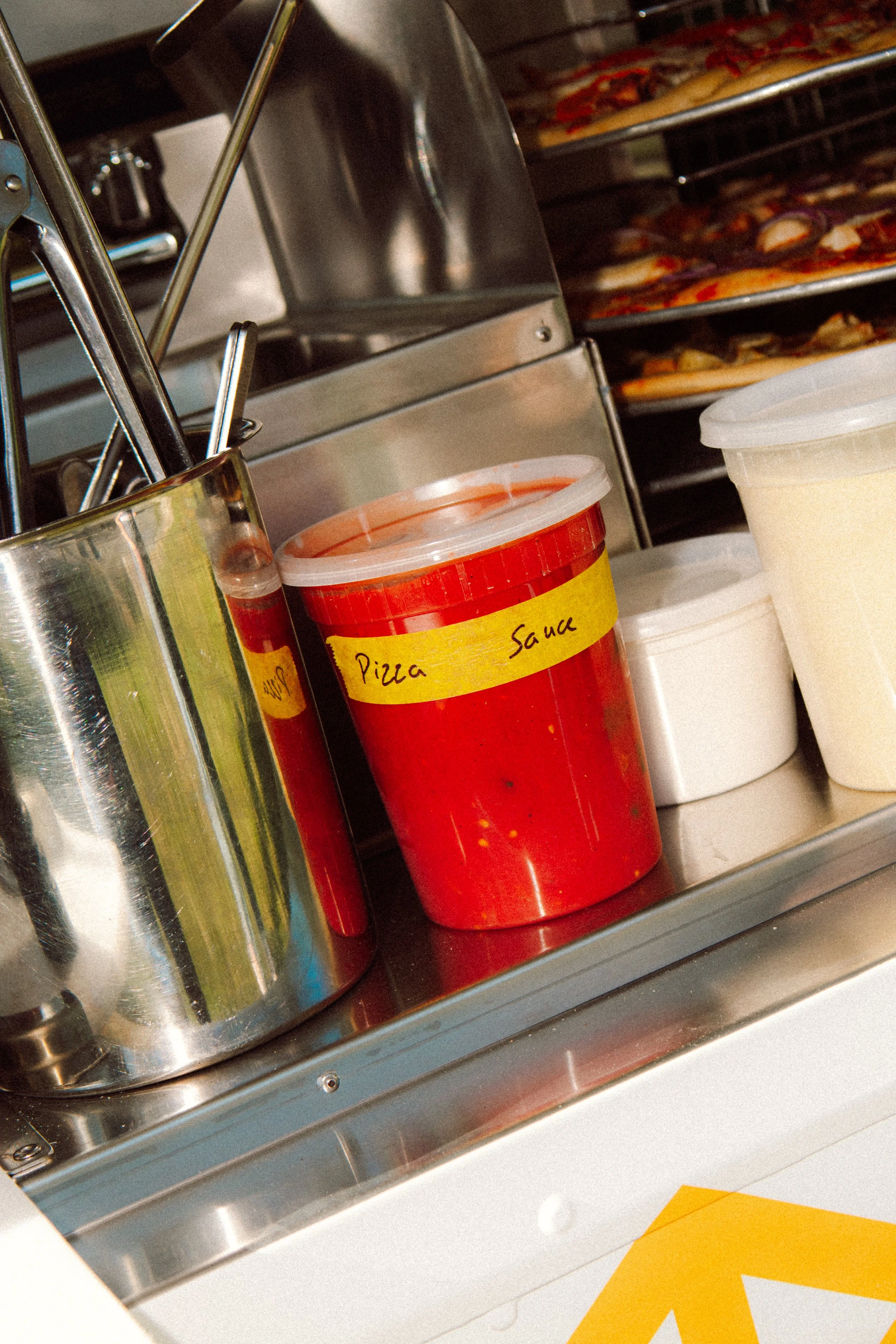 Close-up view of a kitchen or food prep area showing metal containers, a red plastic container labeled 'Pizza Sauce,' and trays of prepared pizzas with toppings such as pepperoni and vegetables, on stainless steel shelves.