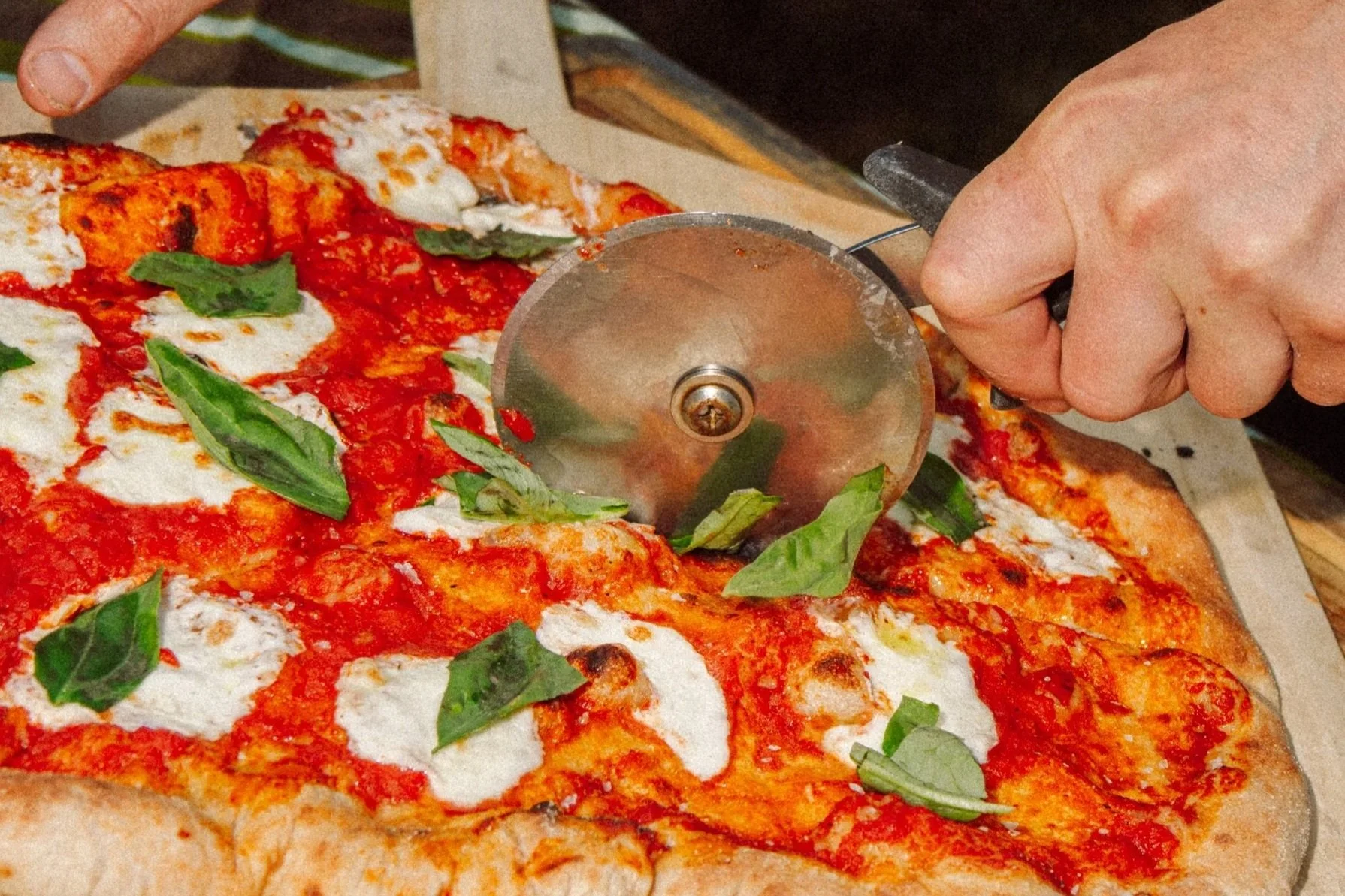 Close-up of a person slicing a freshly baked pizza with basil leaves, melted mozzarella, and tomato sauce on a wooden pizza peel.