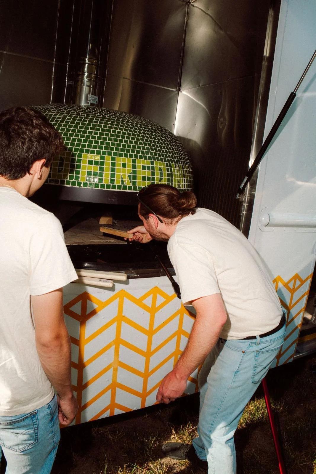 Two men closely inspect a large, colorful mosaic art piece being heated or fired in a kiln or oven, outdoors, with metal walls and a vent pipe visible.