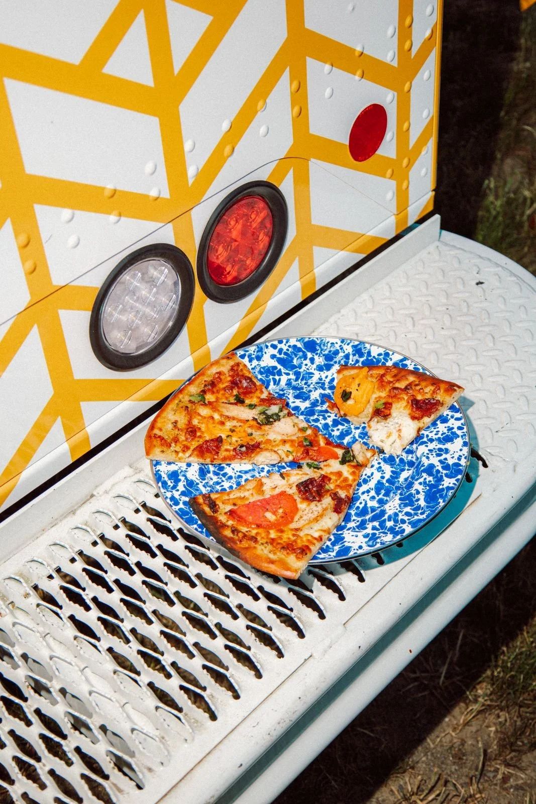 Three slices of pizza on a blue and white plate, placed on a white grill, with a yellow geometric-patterned trailer in the background.