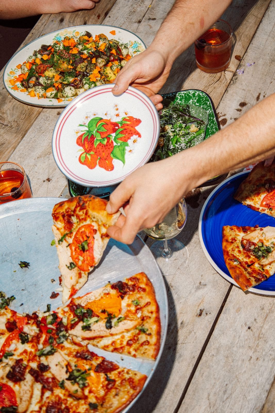 Hands reaching for pizza slices at a table with various dishes, including salad, roasted vegetables, and glasses of drinks.