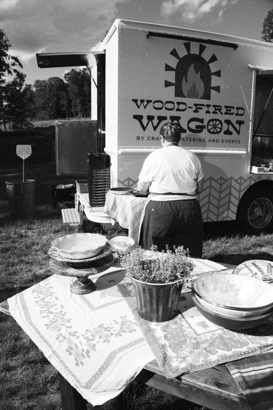A person working at a food truck named "Wood-Fired Wagon" with outdoor tables and bowls set up in front, and a cloudy sky overhead.