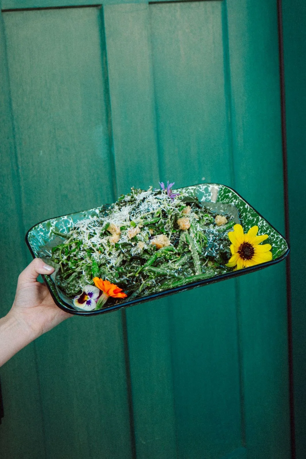 Hand holding a rectangular black bowl filled with a green salad topped with grated cheese and edible flowers, against a green wall background.