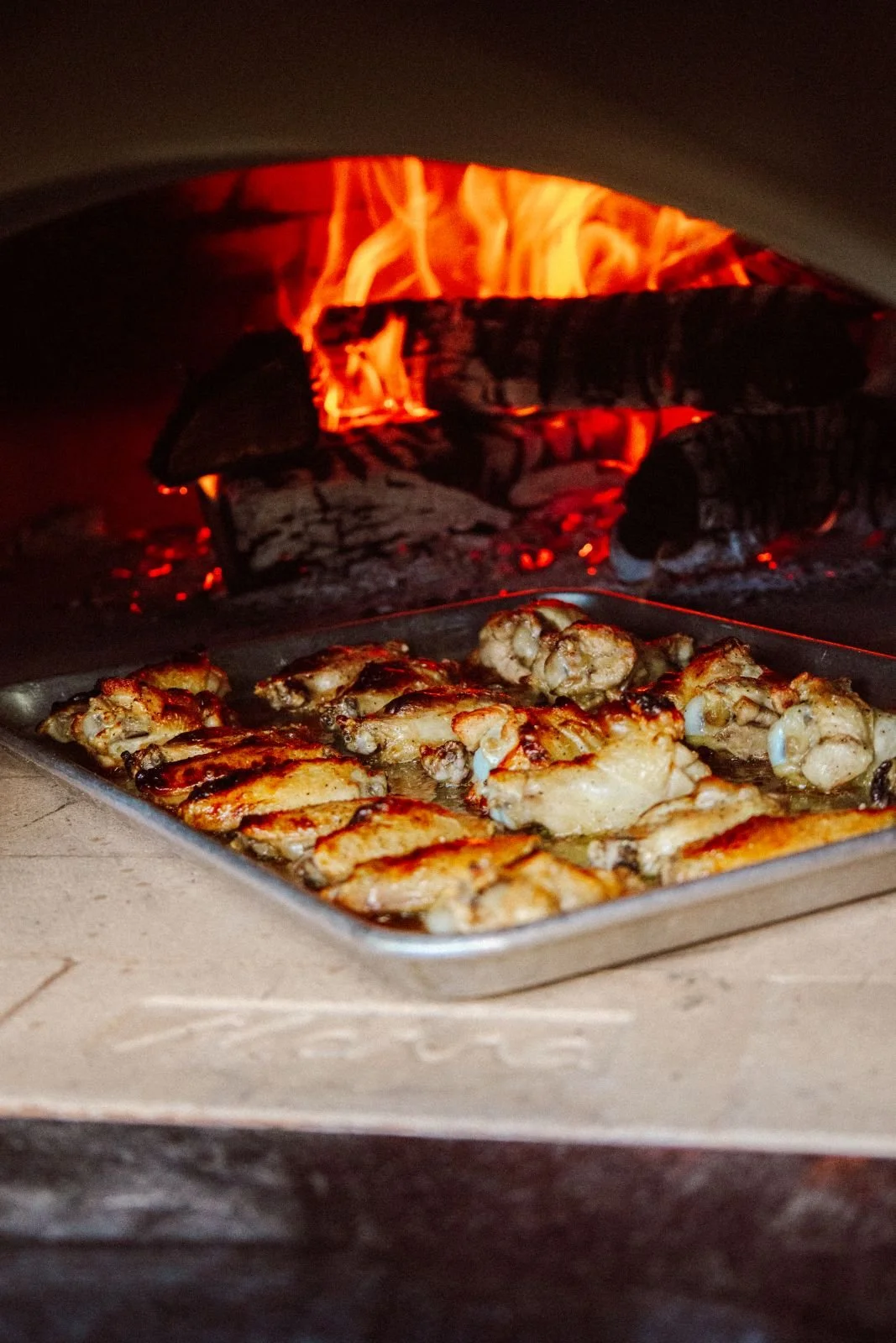 Locally sourced chicken wings baking in a tray in front of a wood-fired oven with flames.