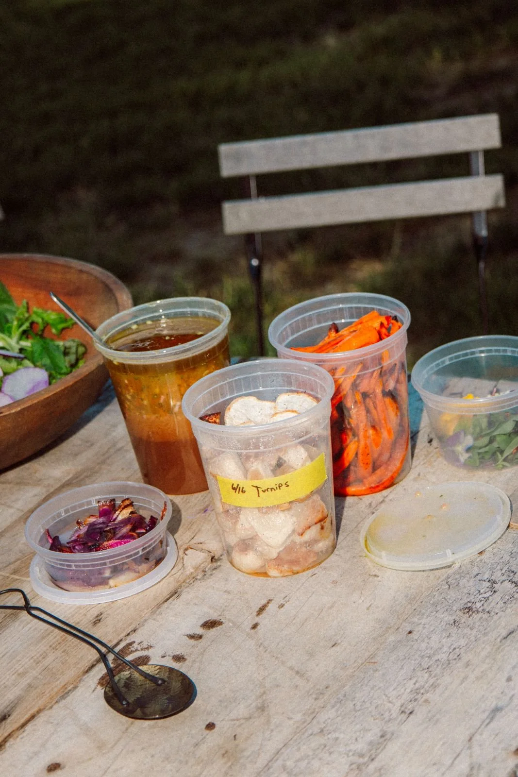 Outdoor table with containers of grilled vegetables, salad, and drinks, set on a rustic wooden surface with a park bench in the background.
