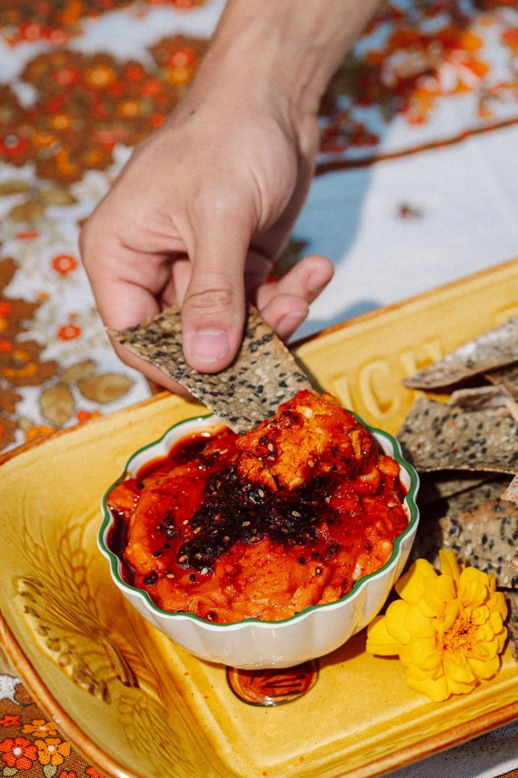 Close-up of hand dipping a black sesame crispbread into a bowl of red chili sauce, with a yellow plate and yellow flower in background.