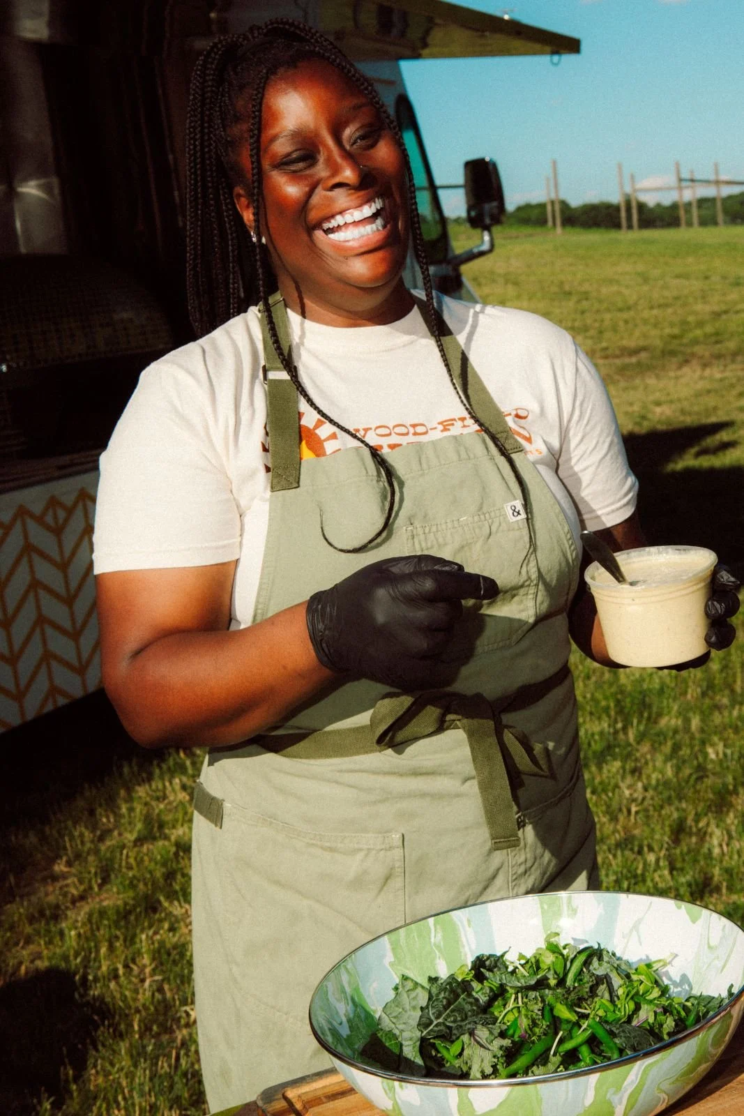 Woman smiling and holding a bowl of salad outdoors, wearing a white t-shirt and an apron, with a grassy field in the background.