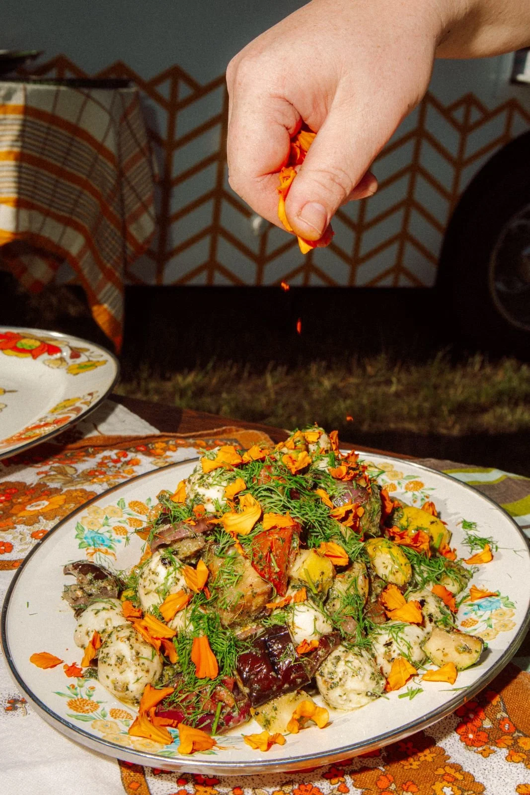 A hand sprinkling orange flower petals over a colorful salad on a decorative plate outdoors.