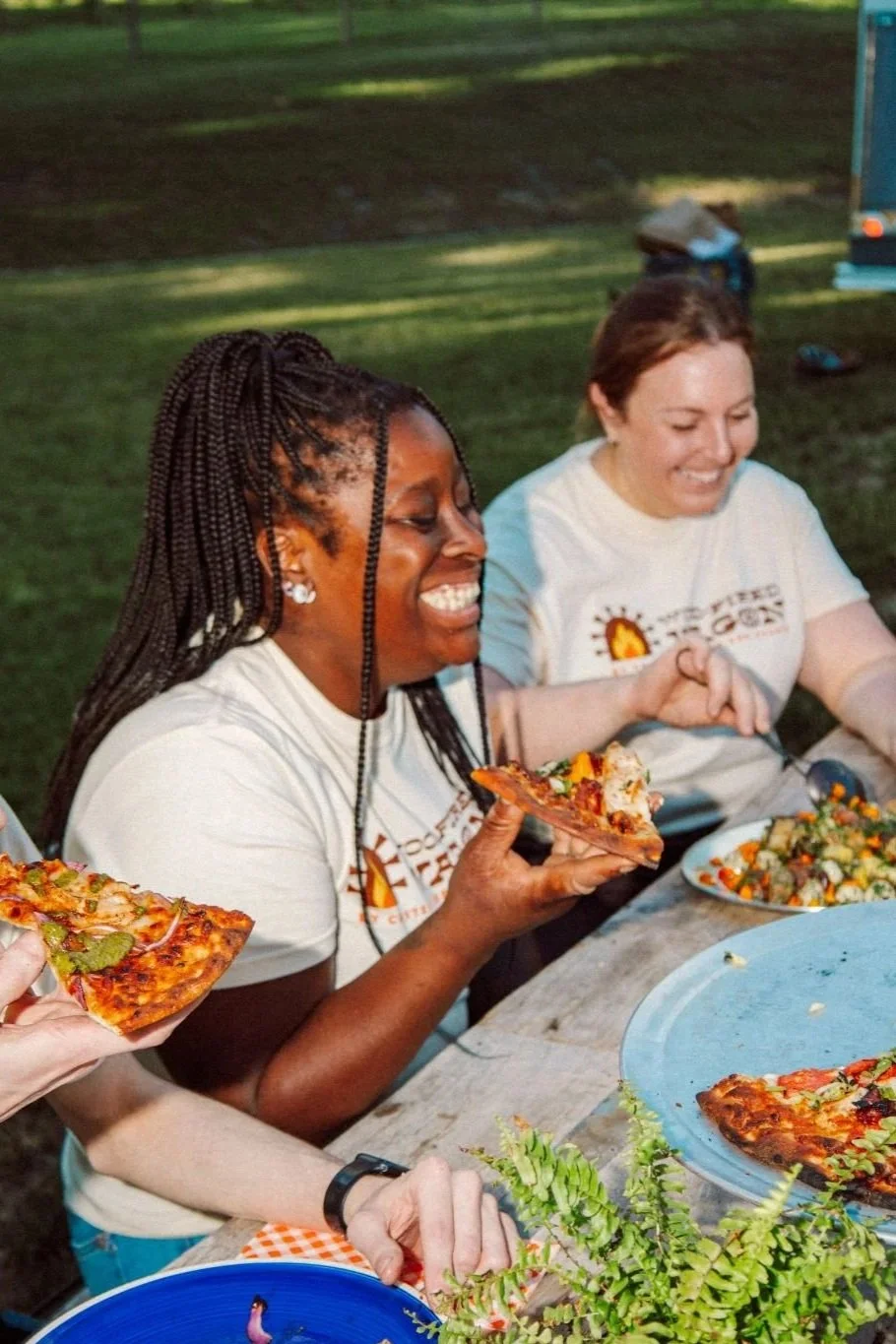 Two women smiling and enjoying pizza at an outdoor gathering, with salads and greenery on the table.