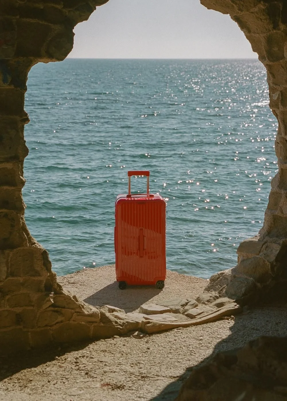 A red suitcase standing on a sandy surface overlooking the ocean, framed by a stone archway.