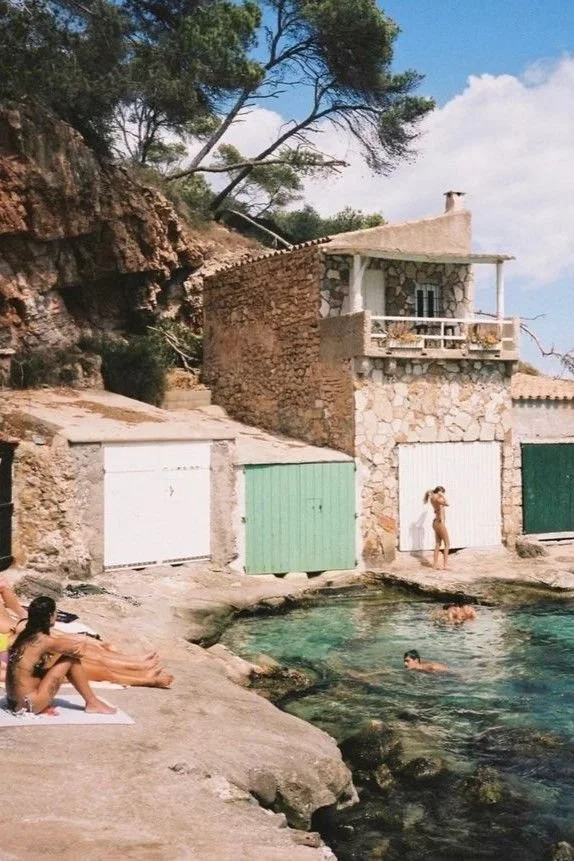 People relaxing and swimming in a small outdoor pool in front of stone houses with colorful garage doors, surrounded by rocky terrain and trees.
