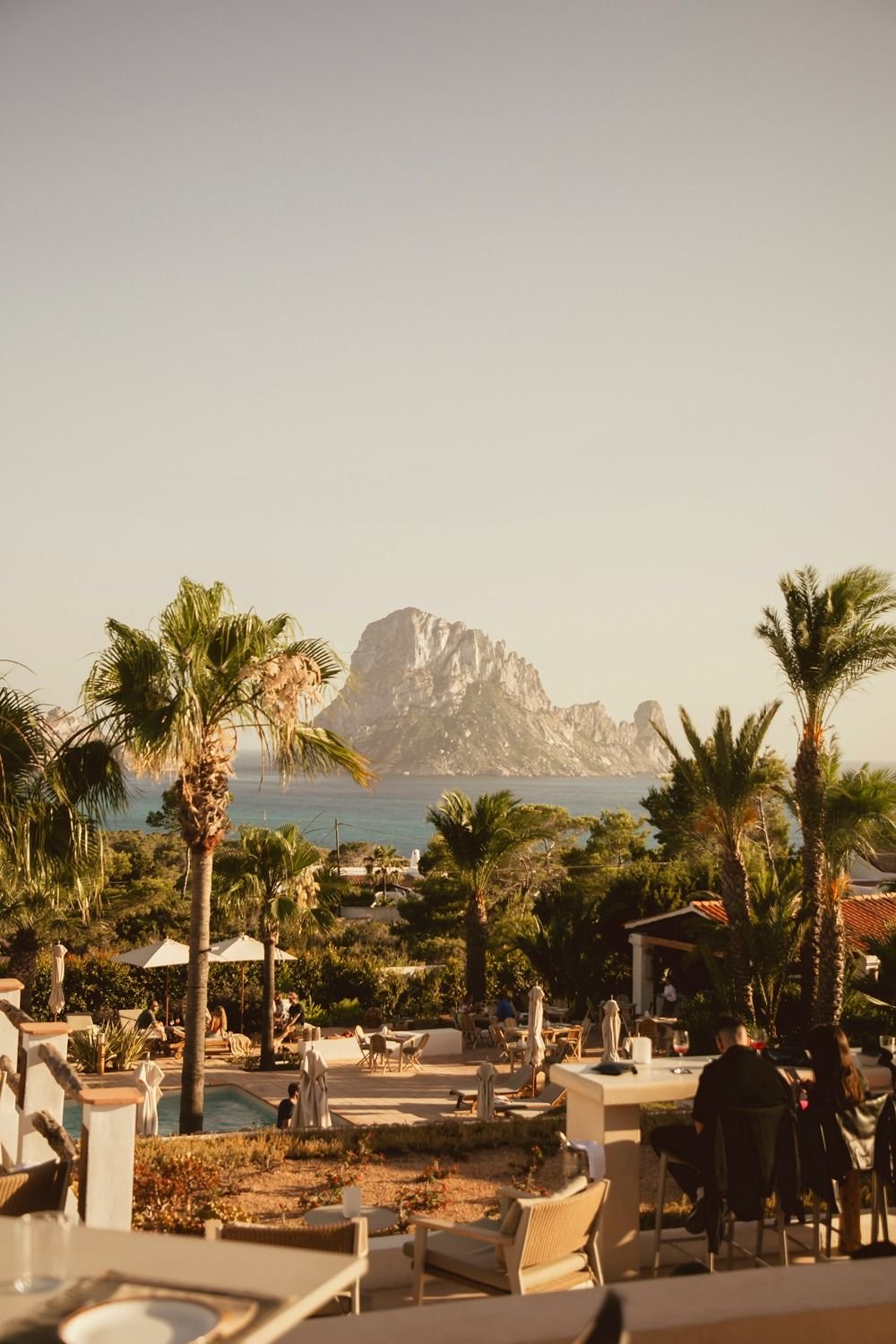 Outdoor restaurant with palm trees, tables, and guests dining, overlooking a mountain and water in the distance during sunset.