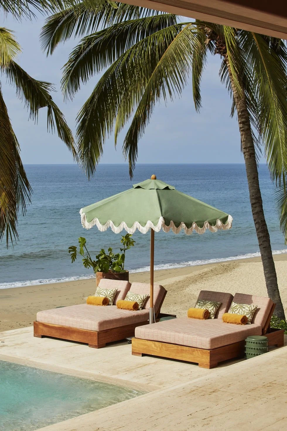 Beachside scene with two lounge chairs under a green umbrella, tropical plants, palm trees, sandy beach, ocean in the background, and part of a pool in the foreground.