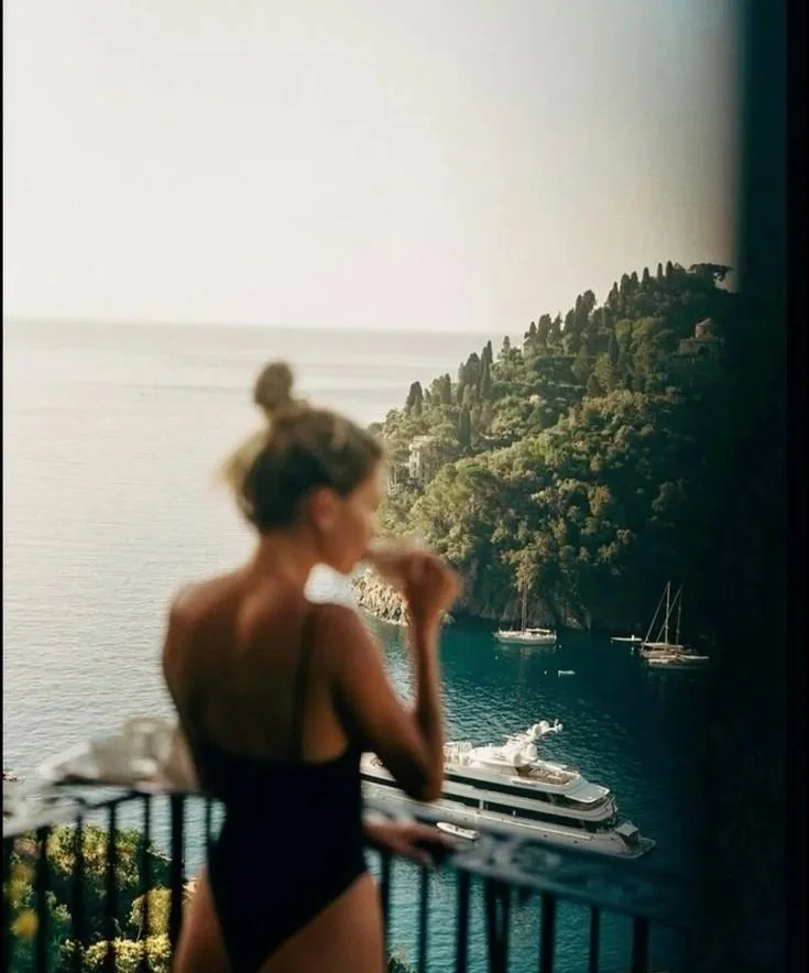 A woman in a black swimsuit standing on a balcony, overlooking a scenic view of boats in a bay, lush green hills, and the ocean.