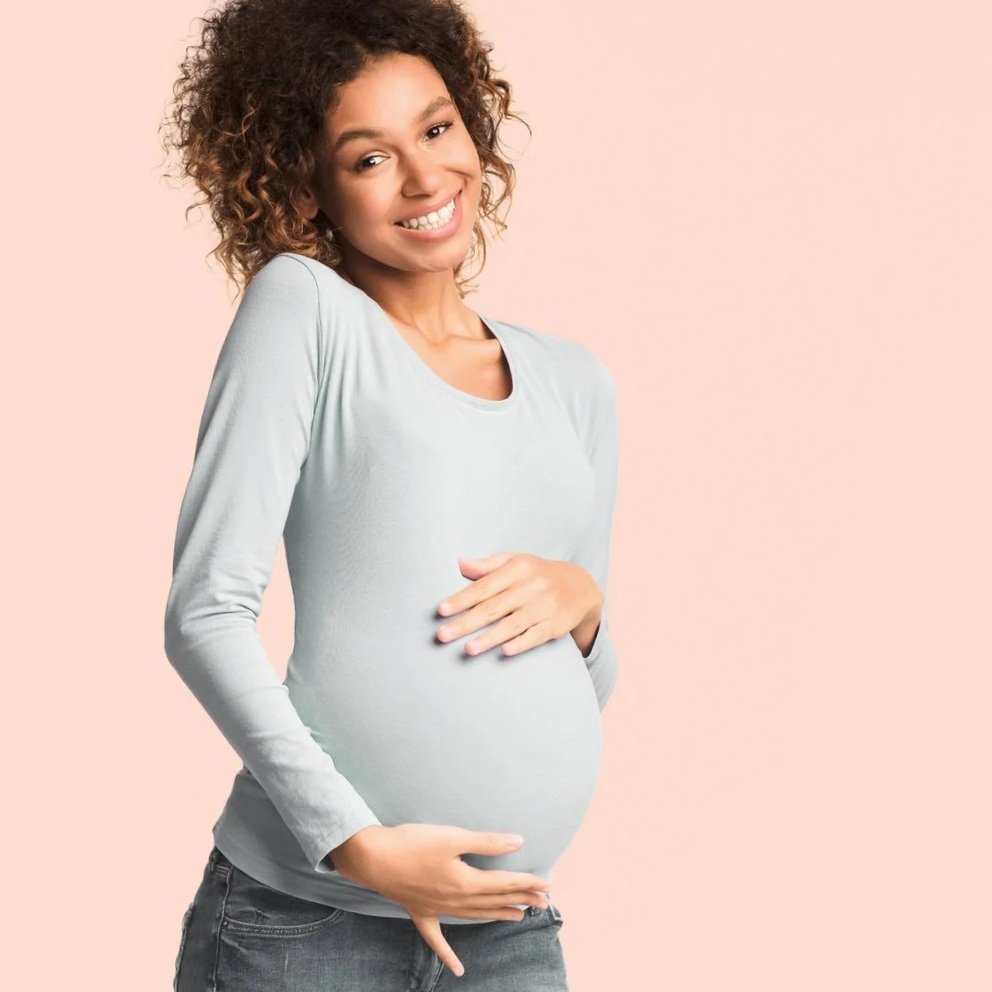 A pregnant woman smiling and touching her belly with one hand, wearing a light gray long sleeve shirt against a pink background.