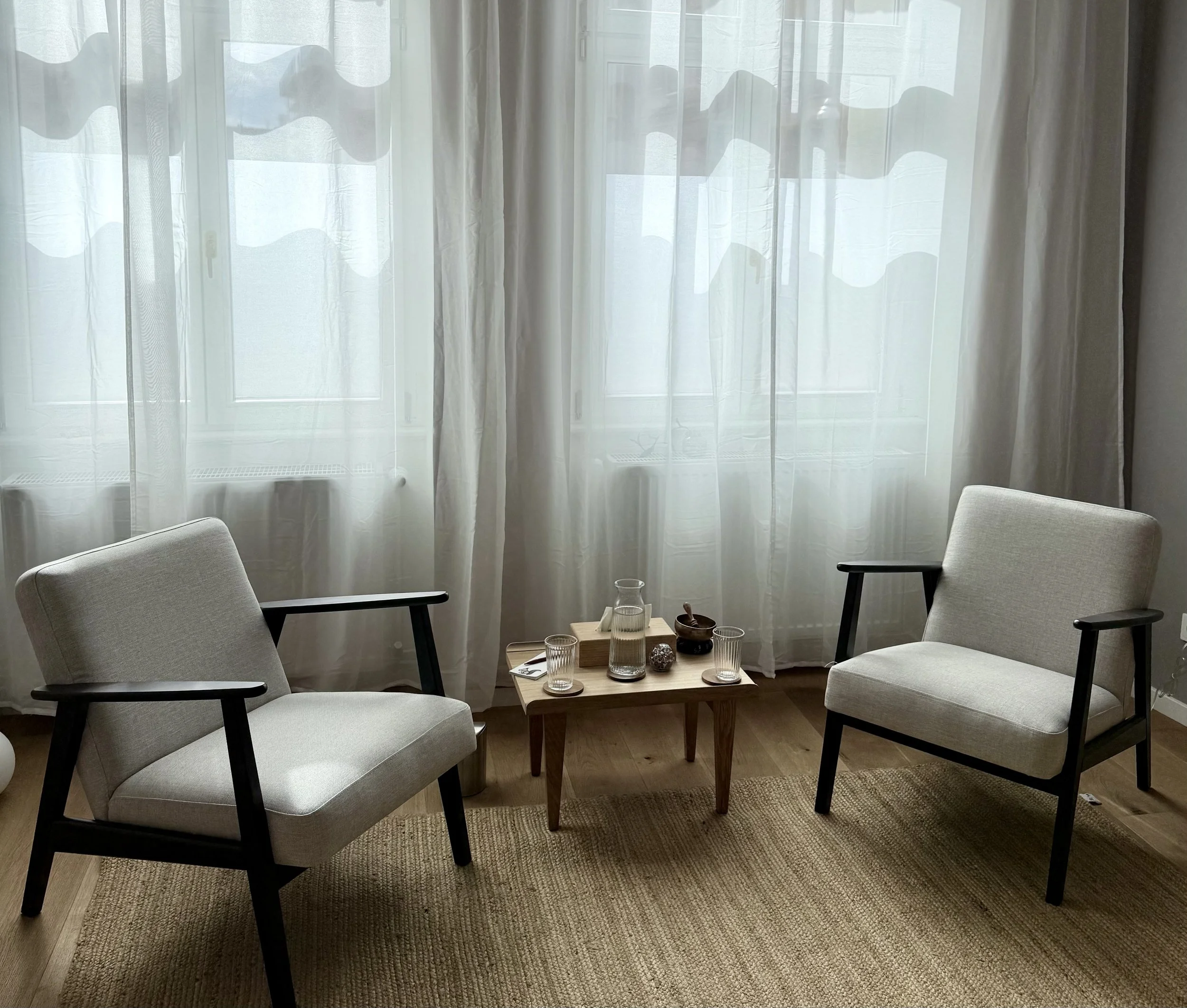 Zwei weiße Sessel mit schwarzen Holzarmen vor einem Fenster mit weißen Vorhängen, zwischen ihnen ein kleiner Holztisch mit Glas Wasser, einigen Gläsern und Dekorationsgegenständen auf einem Teppichboden.