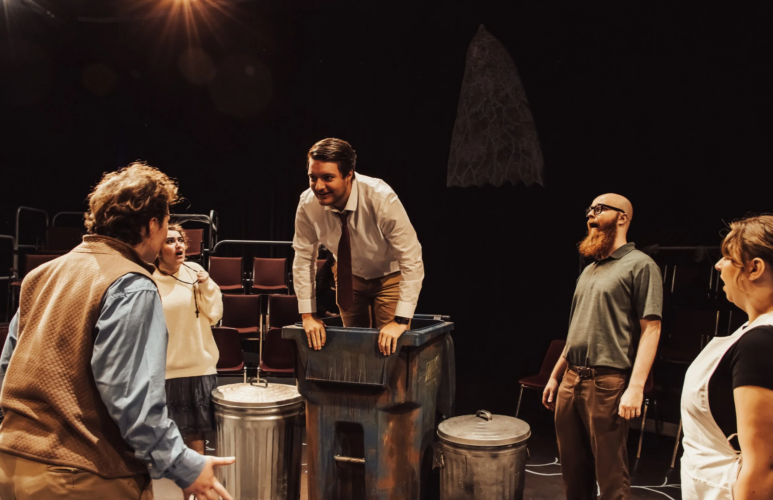 Actors performing a scene on stage, with one actor in a trash bin and others reacting variedly.