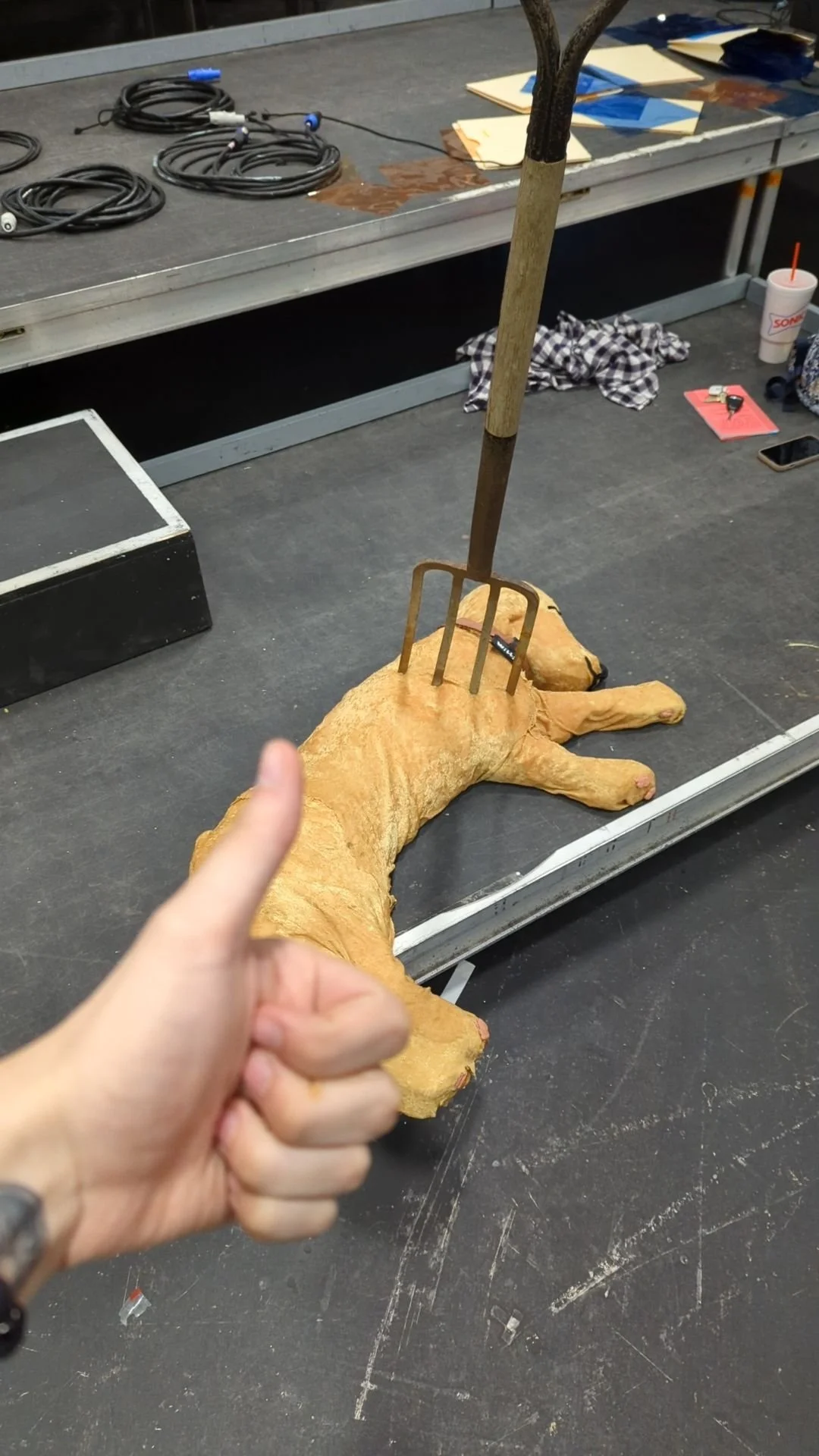 A dog-shaped sculpture made of a material resembling bread, lying on the ground with a garden fork inserted into its back, and a person's hand giving a thumbs-up in the foreground.