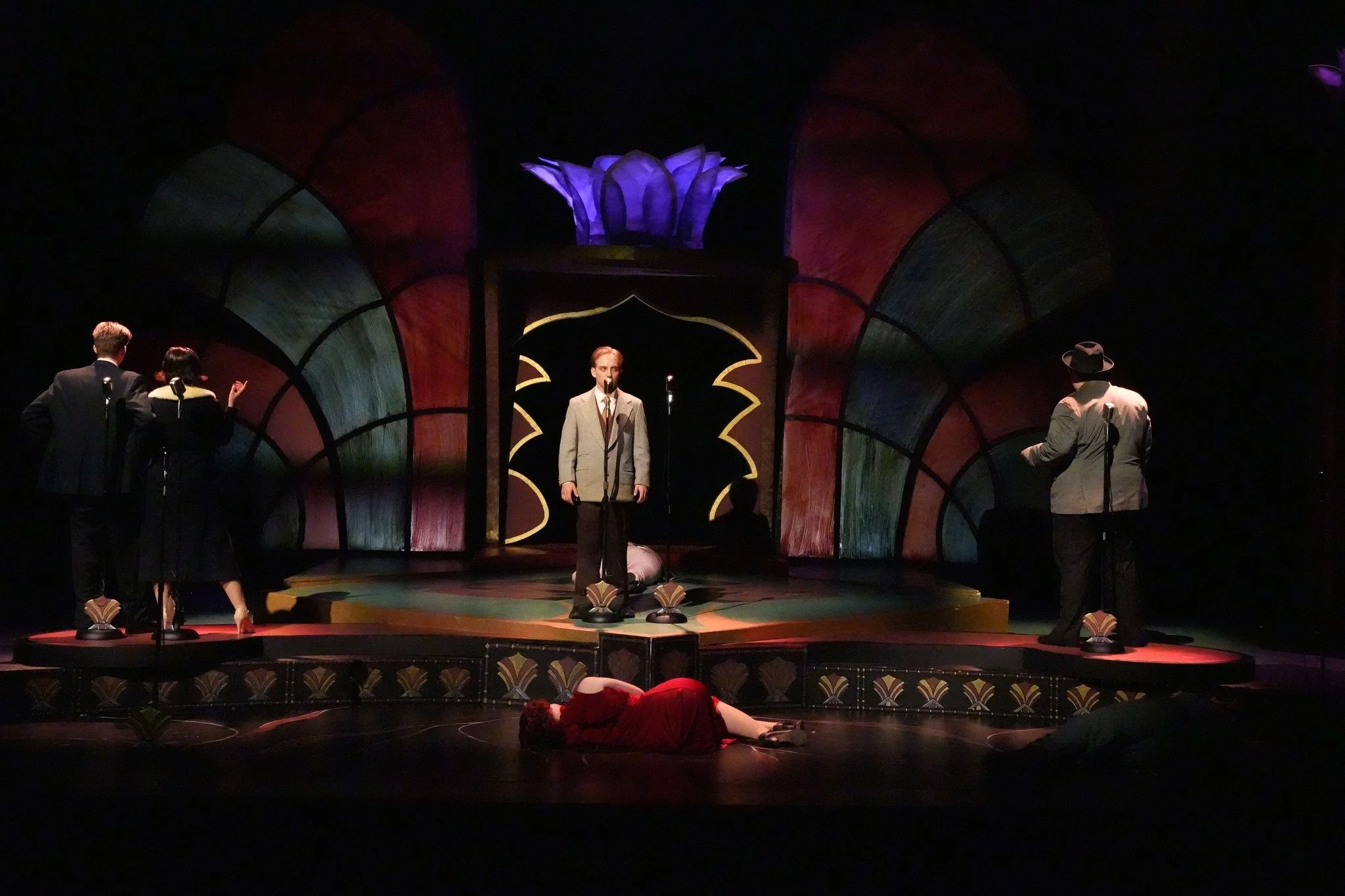 The stage of a theatrical play with four actors; one woman in a red dress lying on the floor in the foreground, and three actors standing in the background, engaged in a scene. The stage has colorful, stained-glass-like arches and a large purple flower prop overhead.