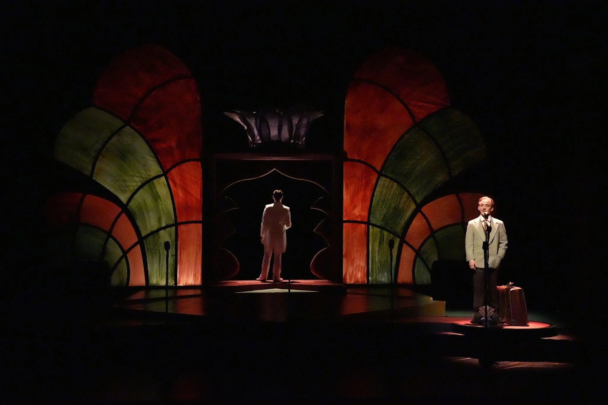 Two performers on a dark stage with stylized, colorful arch designs; one standing with a suitcase and the other facing away in the background.